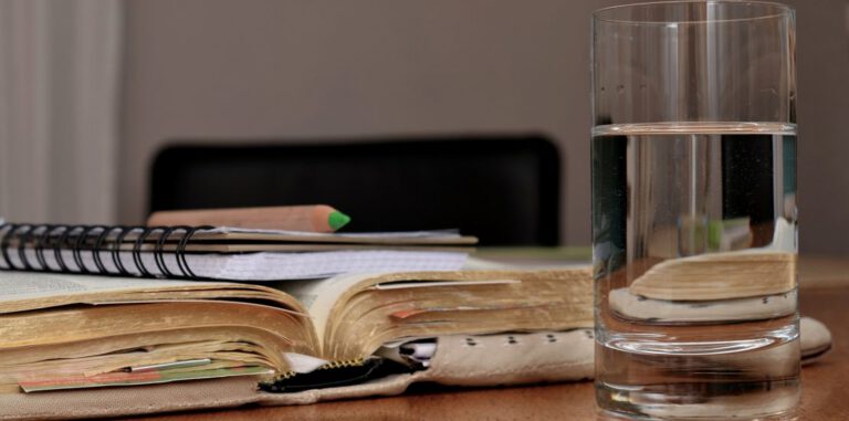Стакан воды на переднем плане и книга с блокнотом и карандашом на заднем фоне.