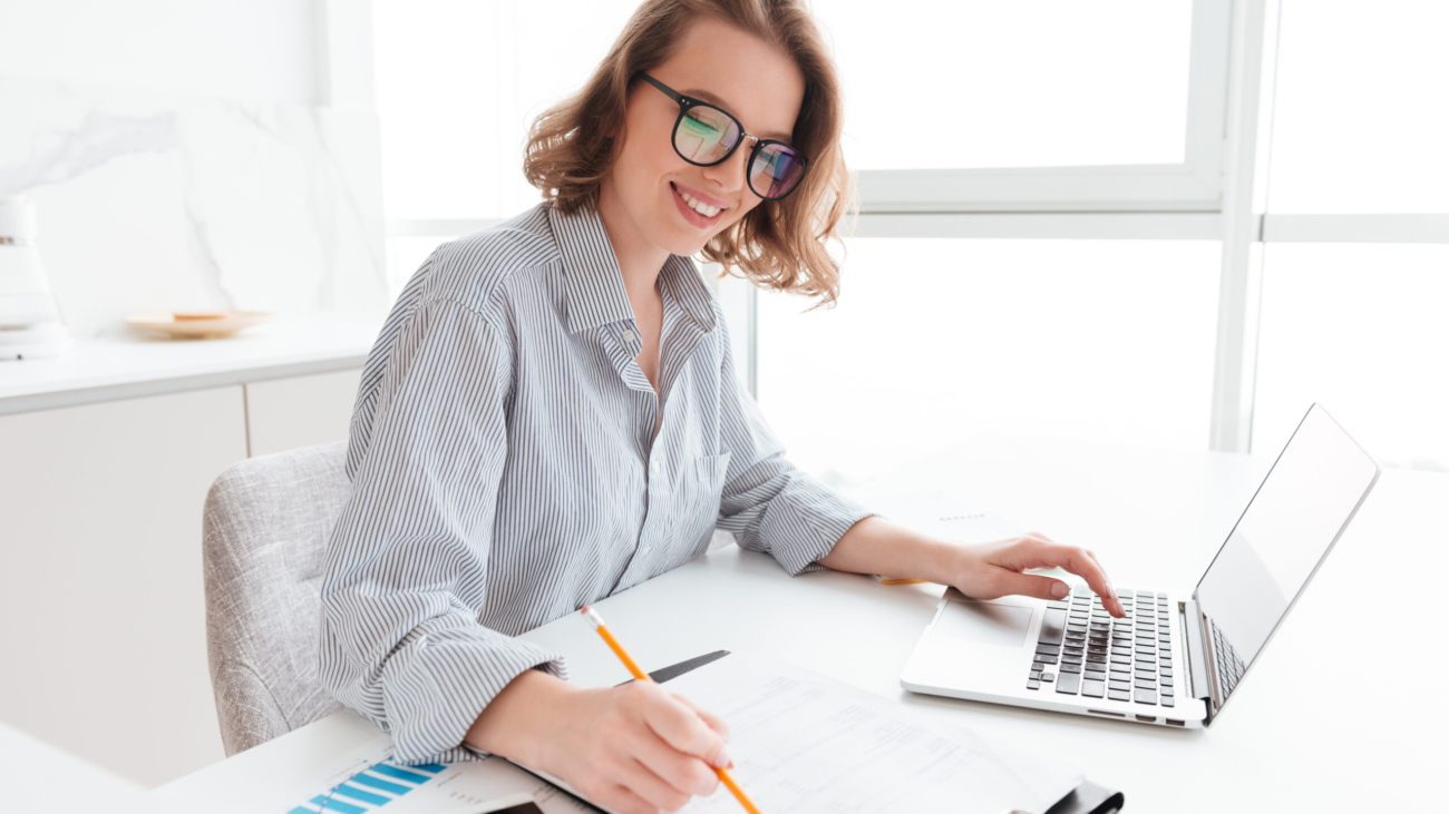 Young attractive smiling girl in glasses and striped shirt working with documents and computer while siting at table in light kitchen