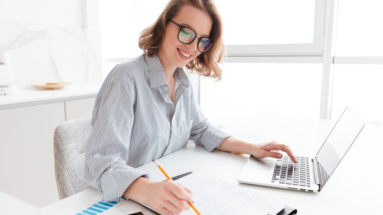 Young attractive smiling girl in glasses and striped shirt working with documents and computer while siting at table in light kitchen