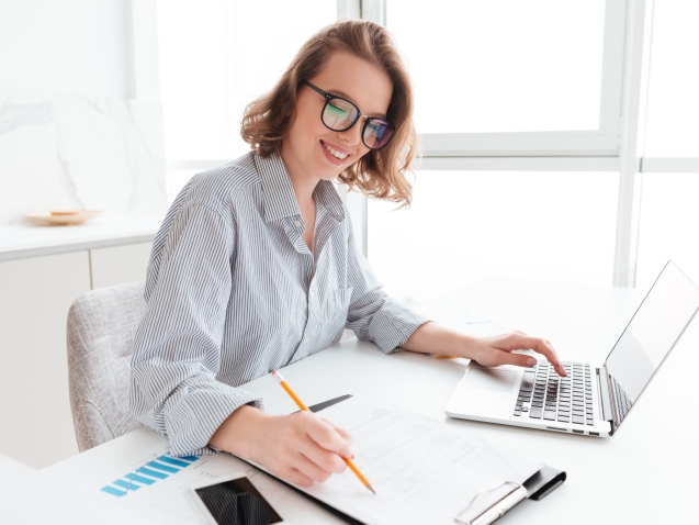 Young attractive smiling girl in glasses and striped shirt working with documents and computer while siting at table in light kitchen
