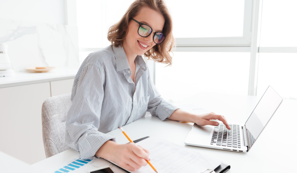 Young attractive smiling girl in glasses and striped shirt working with documents and computer while siting at table in light kitchen